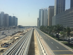 High-rise buildings line a major urban road with separate lanes, bordered by palm trees. Construction work is visible alongside the road with various materials and equipment. The scene is under a clear blue sky, suggesting a sunny day.