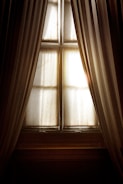 A pair of elegant, long curtains partially opened to reveal a window with diffused sunlight streaming in. The image captures the interplay of light and shadow on soft fabric, creating a warm and serene atmosphere.