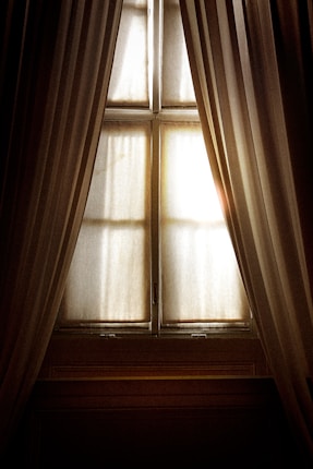 A pair of elegant, long curtains partially opened to reveal a window with diffused sunlight streaming in. The image captures the interplay of light and shadow on soft fabric, creating a warm and serene atmosphere.