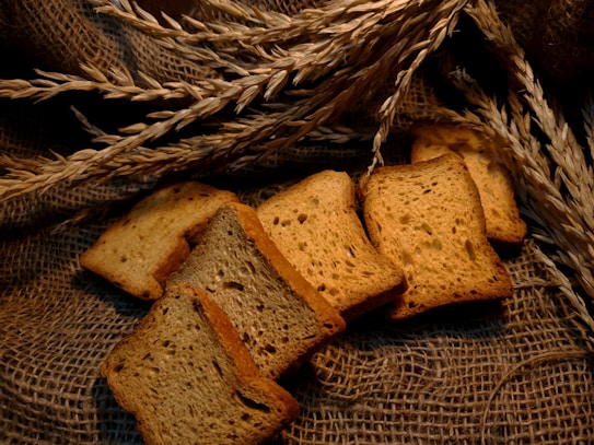 Slices of toasted bread are arranged on a textured burlap background, complemented by sprigs of wheat. The environment gives a rustic and homely feel.