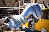 A large, metallic Honda outboard motor is mounted on the stern of a colorful boat. The motor's shiny, silver surface contrasts with the bright yellow and blue hues of the boat. In the background, a rustic stone building adds a textured and historic ambiance to the setting.