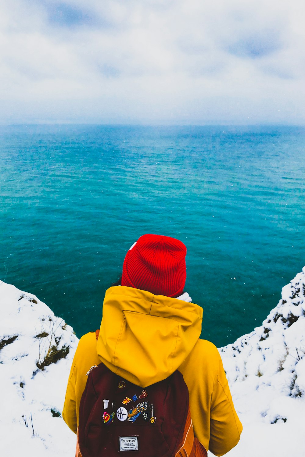 person in yellow hoodie sitting on white snow covered ground near body of water during daytime