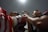 A close-up shot of a boxer preparing for an event with red gloves and a robe featuring star and dragon motifs. Another person is in the background, seemingly assisting or interacting with the boxer. Overhead lighting creates dramatic shadows, emphasizing the intensity of the scene.