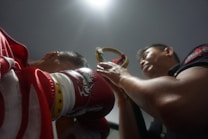 A close-up shot of a boxer preparing for an event with red gloves and a robe featuring star and dragon motifs. Another person is in the background, seemingly assisting or interacting with the boxer. Overhead lighting creates dramatic shadows, emphasizing the intensity of the scene.