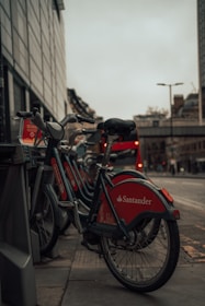 A row of bicycles is parked at a docking station on a city street. The bikes are branded with Santander logos, and their red accents stand out. In the background, a red double-decker bus is partially visible, with urban buildings and a cloudy sky completing the scene.