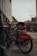 A row of bicycles is parked at a docking station on a city street. The bikes are branded with Santander logos, and their red accents stand out. In the background, a red double-decker bus is partially visible, with urban buildings and a cloudy sky completing the scene.