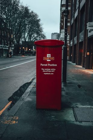A red Royal Mail parcel postbox stands prominently on a city sidewalk. The street appears deserted with buildings lining the road and bare trees on one side, indicating an urban setting. The overall tone is muted, suggesting early morning or evening.