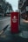 A red Royal Mail parcel postbox stands prominently on a city sidewalk. The street appears deserted with buildings lining the road and bare trees on one side, indicating an urban setting. The overall tone is muted, suggesting early morning or evening.
