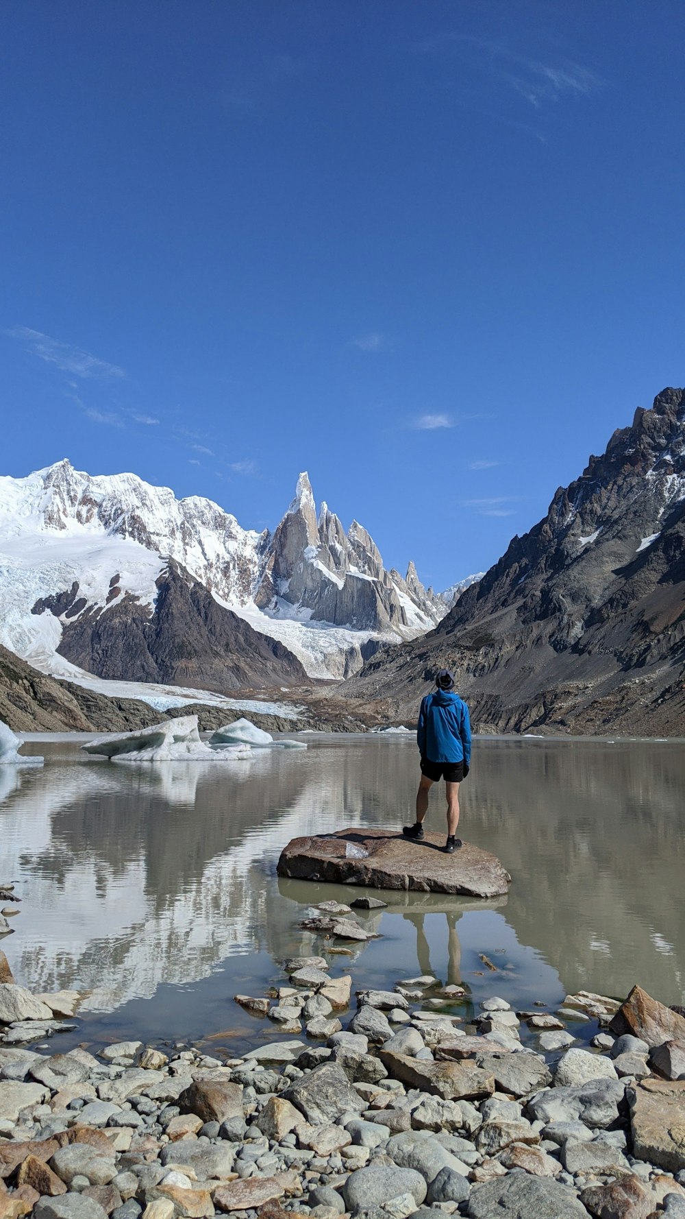 man in blue shirt standing on brown rock near lake during daytime