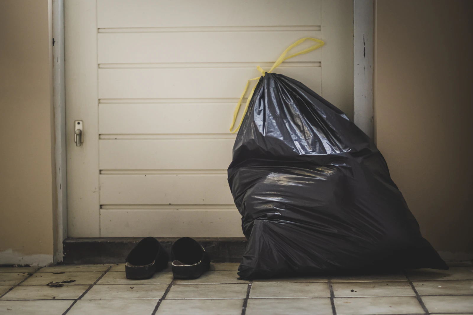 A tied black rubbish bag waiting by a flat's front door, beside a pair of slippers on tiled flooring