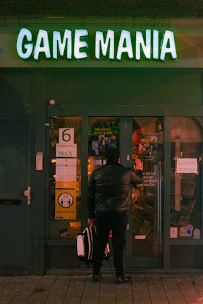 A person dressed in a black leather jacket stands facing a shop named 'Game Mania'. The shop has a glowing green sign and the exterior is dark green. The person is holding a white and black bag and is gazing through the glass door. A sign on the door indicates a maximum of six people allowed inside.