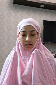 A person is wearing a pink hijab and robe, appearing to be in a contemplative or prayerful state. The background shows a patterned wall and a dark-colored electronic device, possibly a television.