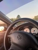 A view from the driver's seat of a car, showing a black steering wheel with the Ram logo. The dashboard in the background has two circular dials with red needles. The side mirror reflects the sunset. Trees and a clear sky are visible through the windshield.