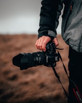 A person is holding a professional camera with a large lens, dressed in a dark and grey jacket. The background is a blurred, earthy landscape, suggesting an outdoor setting.