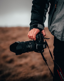 A person is holding a professional camera with a large lens, dressed in a dark and grey jacket. The background is a blurred, earthy landscape, suggesting an outdoor setting.