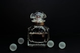 A clear glass bottle of perfume with a rounded cap is positioned against a dark background. The elegant design of the bottle is complemented by its reflective surface, catching subtle highlights. Surrounding the bottle are five frosted glass spheres, evenly dispersed and adding a symmetrical balance to the composition.