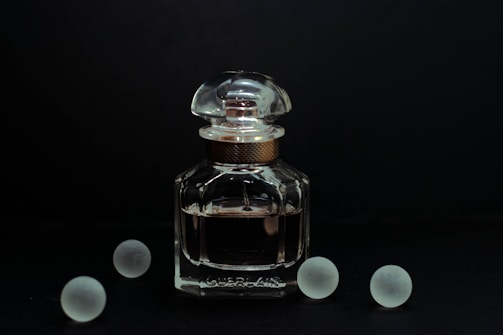 A clear glass bottle of perfume with a rounded cap is positioned against a dark background. The elegant design of the bottle is complemented by its reflective surface, catching subtle highlights. Surrounding the bottle are five frosted glass spheres, evenly dispersed and adding a symmetrical balance to the composition.
