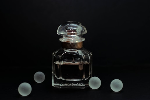 A clear glass bottle of perfume with a rounded cap is positioned against a dark background. The elegant design of the bottle is complemented by its reflective surface, catching subtle highlights. Surrounding the bottle are five frosted glass spheres, evenly dispersed and adding a symmetrical balance to the composition.