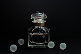 A clear glass bottle of perfume with a rounded cap is positioned against a dark background. The elegant design of the bottle is complemented by its reflective surface, catching subtle highlights. Surrounding the bottle are five frosted glass spheres, evenly dispersed and adding a symmetrical balance to the composition.