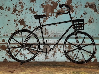 A metal bicycle sculpture is set against a rustic, distressed wall with a textured and speckled pattern in shades of brown and teal. The sculpture features a basket on the handlebars and appears to be made from solid, black metal with a minimalist design.