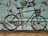 A metal bicycle sculpture is set against a rustic, distressed wall with a textured and speckled pattern in shades of brown and teal. The sculpture features a basket on the handlebars and appears to be made from solid, black metal with a minimalist design.
