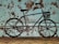 A metal bicycle sculpture is set against a rustic, distressed wall with a textured and speckled pattern in shades of brown and teal. The sculpture features a basket on the handlebars and appears to be made from solid, black metal with a minimalist design.