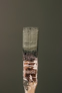 A paintbrush bristling with dark green paint stands vertically against a matching green background. The handle bears signs of wear, with paint splatters and the marking '25MM' visible. The bristles are coated with thick, wet paint.