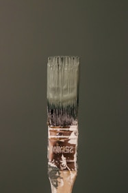 A paintbrush bristling with dark green paint stands vertically against a matching green background. The handle bears signs of wear, with paint splatters and the marking '25MM' visible. The bristles are coated with thick, wet paint.