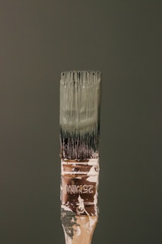 A paintbrush bristling with dark green paint stands vertically against a matching green background. The handle bears signs of wear, with paint splatters and the marking '25MM' visible. The bristles are coated with thick, wet paint.