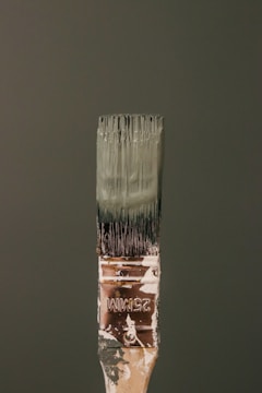 A paintbrush bristling with dark green paint stands vertically against a matching green background. The handle bears signs of wear, with paint splatters and the marking '25MM' visible. The bristles are coated with thick, wet paint.
