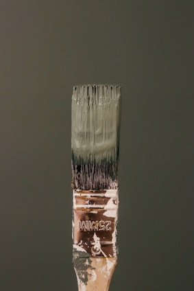 A paintbrush bristling with dark green paint stands vertically against a matching green background. The handle bears signs of wear, with paint splatters and the marking '25MM' visible. The bristles are coated with thick, wet paint.