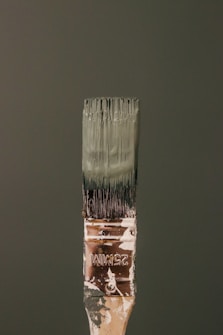 A paintbrush bristling with dark green paint stands vertically against a matching green background. The handle bears signs of wear, with paint splatters and the marking '25MM' visible. The bristles are coated with thick, wet paint.