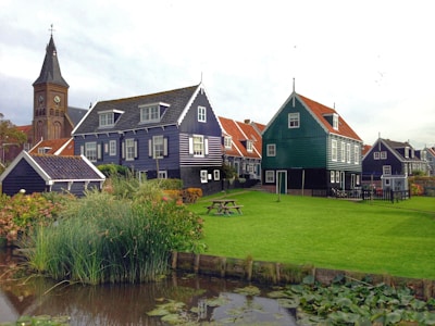 A quaint village scene with traditional Dutch-style houses featuring dark wooden exteriors and orange-tiled roofs. A church with a tall spire is visible in the background. Lush green grass and a neatly maintained garden with a pond are in the foreground, surrounded by reeds and water plants.