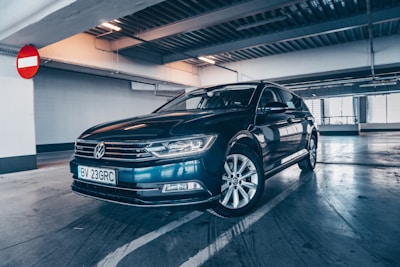 A dark-colored sedan is parked inside a multilevel parking garage. The car is polished and in good condition, with its headlights reflecting the ceiling lights. A no-entry sign is visible on the wall behind the vehicle, and the concrete floor shows some markings.