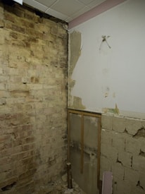 A partially unfinished room with exposed brick walls and a section of plaster that has been chipped away. The ceiling has white tiles, and some construction debris is visible on the floor.