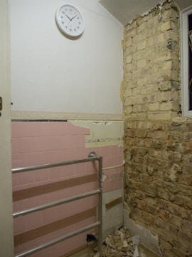 A partially renovated room with a tiled pink wall featuring some missing tiles, a metal railing, exposed brick on one wall, and a basic round wall clock showing the time as approximately 10:10. Debris is scattered on the floor, indicating ongoing construction or renovation work.
