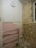 A partially renovated room with a tiled pink wall featuring some missing tiles, a metal railing, exposed brick on one wall, and a basic round wall clock showing the time as approximately 10:10. Debris is scattered on the floor, indicating ongoing construction or renovation work.