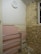 A partially renovated room with a tiled pink wall featuring some missing tiles, a metal railing, exposed brick on one wall, and a basic round wall clock showing the time as approximately 10:10. Debris is scattered on the floor, indicating ongoing construction or renovation work.