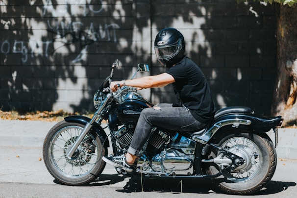 Biker wearing a matte black helmet and gloves, standing next to a roaring motorcycle on a sunny day.