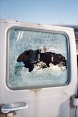 A close-up view of a car door with a shattered window. The interior of the vehicle is visible through the broken glass, displaying damaged or disarrayed upholstery. The scene is set against a clear blue sky, suggesting an outdoor environment.