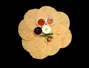A collection of circular, flatbreads arranged in a flower-like pattern on a black background. In the center, there are small bowls containing various condiments such as red and green chutneys, yogurt, and sliced vegetables including onions and cucumbers. There are also whole green chilies and a red onion placed on top of the flatbreads.