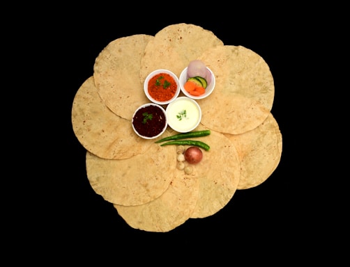 A collection of circular, flatbreads arranged in a flower-like pattern on a black background. In the center, there are small bowls containing various condiments such as red and green chutneys, yogurt, and sliced vegetables including onions and cucumbers. There are also whole green chilies and a red onion placed on top of the flatbreads.