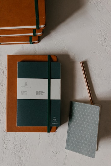Several neatly stacked brown notebooks with green elastic bands and a calendar notebook with a pen on a textured light gray surface.
