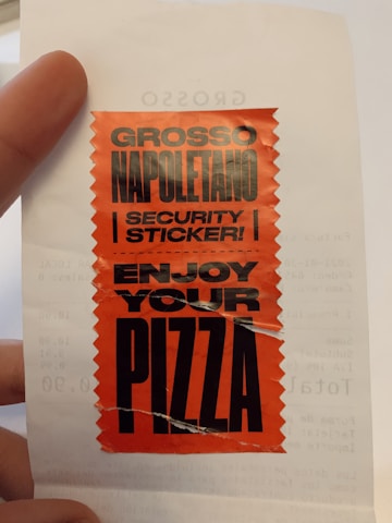 A red sticker with a jagged design borders, featuring bold black text that reads 'GROSSO NAPOLETANO SECURITY STICKER! ENJOY YOUR PIZZA'. The sticker is placed on a white piece of paper, possibly part of a receipt. A person's hand is partially visible holding the paper.