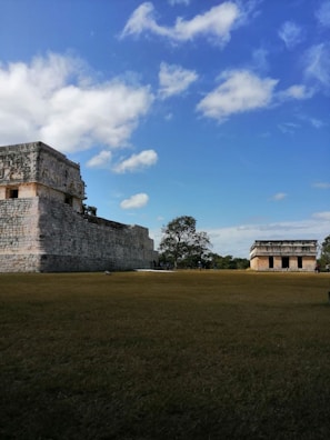 Ancient stone structures stand on a grassy field under a bright blue sky with scattered white clouds. There are two main buildings, one larger with detailed stone carvings and another smaller in the background.