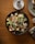 A bowl of fresh salad containing lettuce, croutons, grilled chicken pieces, cherry tomatoes, grated cheese, and microgreens, all lightly drizzled with creamy dressing. The salad is presented in a dark bowl placed on a wooden table, with a small white plate holding a salt shaker nearby and a partial view of another plate.
