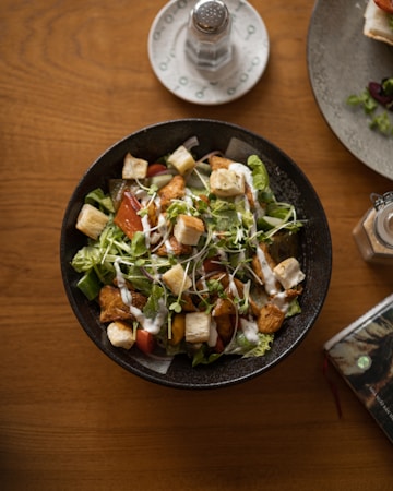 A bowl of fresh salad containing lettuce, croutons, grilled chicken pieces, cherry tomatoes, grated cheese, and microgreens, all lightly drizzled with creamy dressing. The salad is presented in a dark bowl placed on a wooden table, with a small white plate holding a salt shaker nearby and a partial view of another plate.