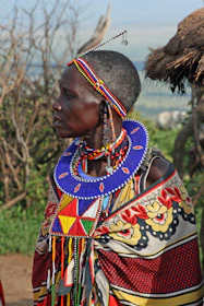 A Maasai warrior greeting visitors near a traditional village under a bright blue sky.