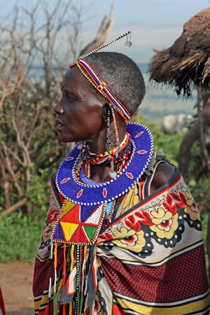A person adorned in traditional, colorful Maasai attire stands outdoors. The individual wears a large blue beaded necklace, beaded headband, and other intricate beadwork. There is a shawl draped over the shoulders featuring bold floral patterns and vibrant colors such as red, yellow, and black. In the background, there are blurred elements like green foliage and a thatched structure.