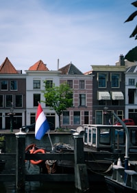 A serene canal scene featuring traditional Dutch buildings with triangular rooftops, a tree, and a boat docked in the foreground. A flag with red, white, and blue stripes is prominently displayed on the boat, and several lifebuoys hang on posts.